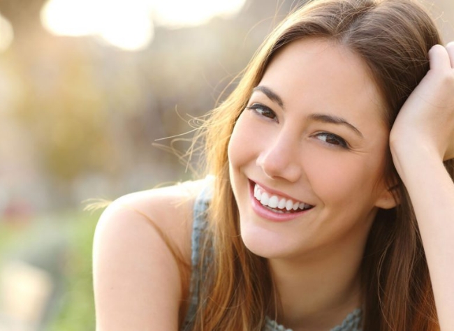 Smiling woman with long brown hair, resting her head on her hand outdoors in soft natural light.